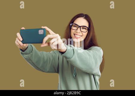 Glückliche junge Frau mit Hoodie und Brille lächelt und macht Selfie auf ihrem Handy Stockfoto