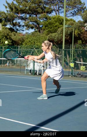 Selbstbewusste junge kaukasische Tennisspielerin trifft am sonnigen Tag mit Schläger auf dem Platz den Ball Stockfoto