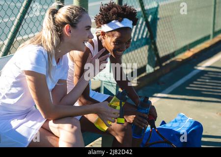 Glückliche junge, multirassische Tennisspielerinnen genießen am sonnigen Tag eine Pause, während sie auf dem Platz reden Stockfoto