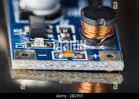 Elektronische Spule, schwarzes DC/DC-Wandlermodul, LED-Diode oder Kondensator auf blauem Leiterplattendetail mit Reflexion auf glänzendem schwarzem Hintergrund. SMD-Leiterplatte. Stockfoto