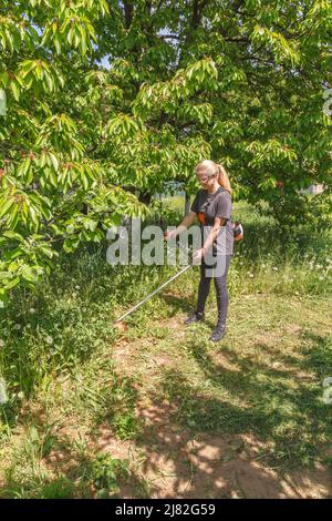 Eine Frau mäht das Gras im Garten mit einem Benzintrimmer. Sie trug eine Brille, um seine Augen zu schützen. Arbeiten Sie im Frühling im Garten. Stockfoto