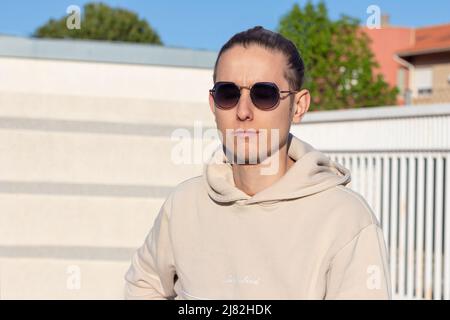 Porträt eines jungen Mannes in einem beigen Sweatshirt und einer schwarzen Sonnenbrille an einem Frühlingstag, der in die vordere Sonne blickt Stockfoto