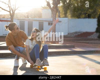 Heterosexuelles junges Paar, das eine gute Zeit hat, während es ein Longboard im Nachmittagslicht wie Kinder benutzt Stockfoto