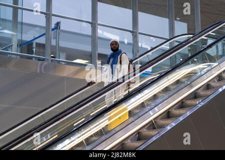 Afroamerikanischer Passagiermann mit Koffer steht auf Rolltreppe, hält Handlauf im Flughafenterminal Stockfoto