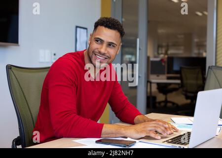 Glücklicher hispanischer Berater, der mit einem Laptop im Sitzungssaal am modernen Arbeitsplatz sitzt und wegschaut Stockfoto