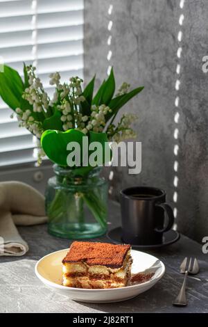 Traditionelles italienisches Dessert Tiramisu, Kaffee und ein Bouquet von Lilien des Tals auf grauem Betongrund. Stockfoto