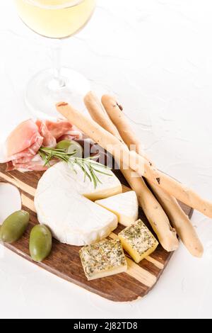 Holzbrettaufschnitt mit einer Auswahl an Käse und Prosciutto, serviert mit Oliven, Grissini und einem Glas Wein auf einem Tisch aus weißem Stein. Italienische Traditionti Stockfoto