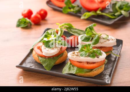 Bruschettas mit Caprese-Salat, geschnittenen Tomaten, Mozzarella, Basilikum und gekrönt mit Mais-Salat auf schwarzem Teller, Holztisch Hintergrund. Klassisches italienisch Stockfoto