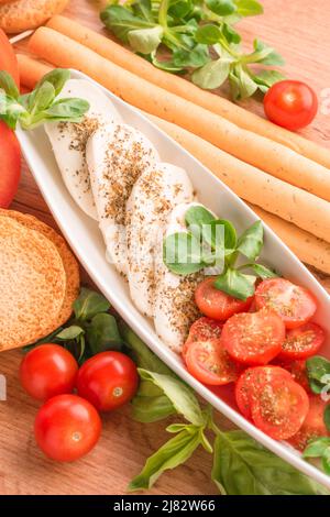 Vertikales Bild des traditionellen italienischen Caprese-Salats mit Babytomaten und Mozzarella in Scheiben, Basilikum, Olivenöl, Maissalat, Bruschettas und Grissini o Stockfoto