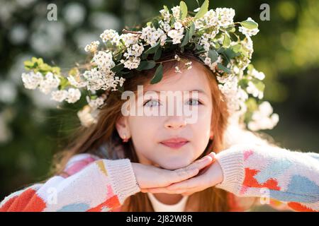 Glücklich lächelnd Kind Mädchen 6-7 Jahre alt tragen Strick bunten Pullover und Blumenkranz Frisur mit Blumen im Park über Natur Hintergrund. Frühling. Sp Stockfoto