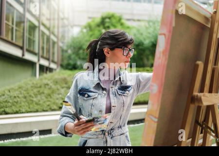 Junge Künstlerin keuchend Leinwand auf Staffelei im Freien in Park Stadt - Fokus auf Maler Frau Gesicht Stockfoto