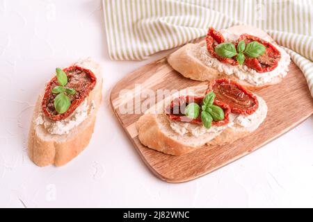 Bruschettas mit Ricotta-Käse, sonnengetrocknete Tomaten in Scheiben geschnitten, mit Basilikum auf einem hölzernen Servierbrett, weißer Steintisch im Hintergrund. Klassisches italienisches und Stockfoto