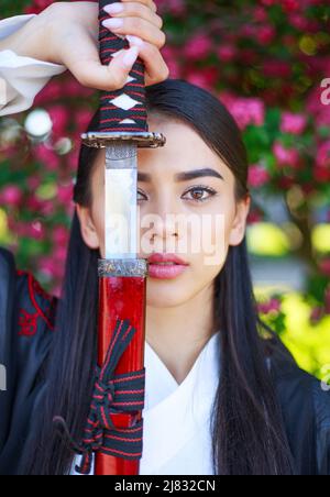 Elegantes Mädchen in einem traditionellen Kimono in einem blühenden Blumengarten mit einem Samurai japanischen Schwert Katana in das Bild einer asiatischen Kriegerin Stockfoto