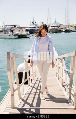 Die ganze Länge der lächelnden Frau in blauem Hemd und weißen Hosen, die auf dem Pier im Hafen spazieren gehen Stockfoto