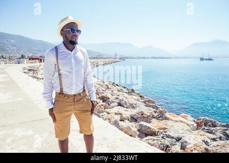 Hübscher afroamerikanischer Mensch lässiger Mode Look Outfit in Strohhut, weißes Hemd zu Fuß durch Tyrkey Küste Stockfoto