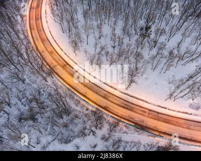 Fliegen Sie über Winter schneebedeckten leeren Straßenverlauf. Beleuchtete abendliche Autobahn, umgeben von schneebedecktem Wald. Luftaufnahme nach unten Stockfoto