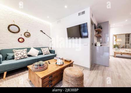 Apartment mit offener Küche und Wohnzimmer mit sichtbaren Betonsäulen, dreisitziger blauer Stoffsessel, an der Wand montiertem TV und Vintage-Beistelltisch Stockfoto