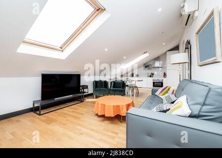 Penthouse-Apartment mit offener Küche und Wohnzimmer, blauem Stoff-Klappsofa für 3 Personen, TV auf schwarzem Tisch und Vintage-Beistelltisch, Parkett f Stockfoto