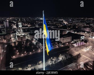 Beleuchtete Flagge der Ukraine winkt über dem Stadtbild der dunklen Nacht. City Center Luftaufnahme in der Nähe der Dormition Kathedrale in Kharkiv, Ukraine. Farblos Stockfoto