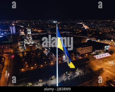 Beleuchtete Flagge der Ukraine winkt über dem Stadtbild der dunklen Nacht. City Center Luftaufnahme in der Nähe der Dormition Kathedrale in Kharkiv, Ukraine Stockfoto