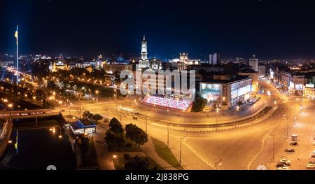 Beleuchteter Flussdamm mit Flagge der Ukraine und Wasserspiegelung in der Nacht. Stadtpanorama über dem Fluss Lopan in der Nähe der Kathedrale von Dormition Stockfoto