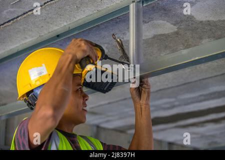 Facharbeiter im Bauwesen, der eine C-Klemme und einen elektrischen Schraubendreher verwendet, um eine Stahlstrebe an einer Stützstrebe für eine abgehängte Decke in einem zu befestigen Stockfoto