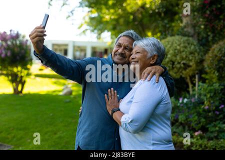 Lächelnder älterer Biracial-Mann, der im Park stehend mit seiner Frau ein Selfie über das Mobiltelefon gemacht hat Stockfoto