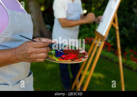 Mittelteil der Biracial Senior Frau hält Palette und Pinsel, während Mann malen im Hintergrund Stockfoto