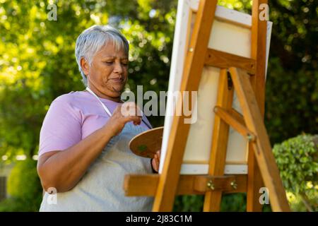 Low-Angle-Ansicht der Biracial Senior Frau mit kurzen Haaren Malerei mit Aquarellen auf Leinwand im Hof Stockfoto