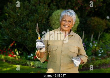 Porträt einer lächelnden älteren Frau aus der Birazialzeit mit kurzen Haaren, die Handschuhe trägt und die Gartengabel hält Stockfoto