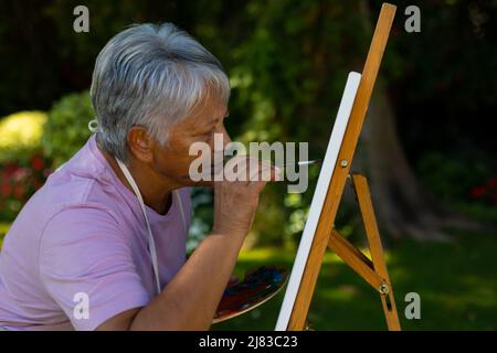 Fokussierte ernsthafte Biracial ältere Frau mit kurzen Haaren Malerei auf Leinwand mit Aquarellen im Hof Stockfoto