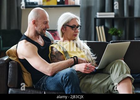Stilvoller, reifer kaukasischer Mann und verliebte Frau entspannen sich auf der Couch im Wohnzimmer zu Hause und schauen sich Filme im Internet auf dem Laptop an Stockfoto