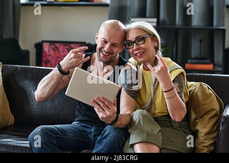 Fröhliches reifes Paar in legerer Kleidung auf dem Sofa im Wohnzimmer sitzen und Spaß beim Fotografieren von Selfie-Bildern auf einem digitalen Tablet haben Stockfoto