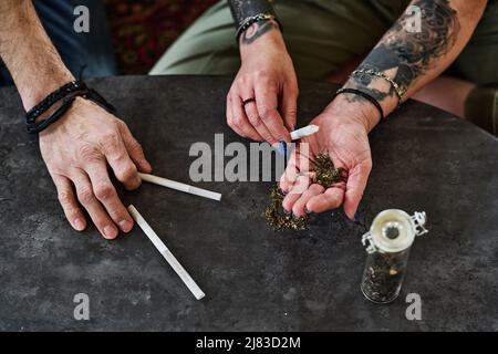 Blick aus der Perspektive auf unverkennbare reife Männer und Frauen, die am Tisch sitzen und Cannabis-Zigaretten machen Stockfoto