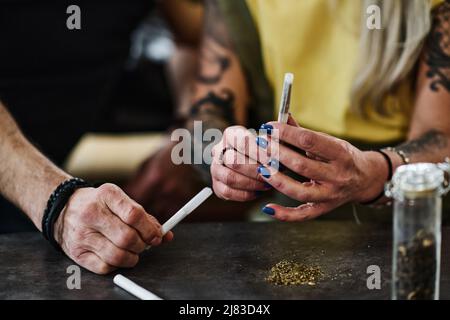 Nicht erkennbare reife tätowierte Frau und ihr Freund sitzen mit Marihuana an Tischrollgelenken Stockfoto