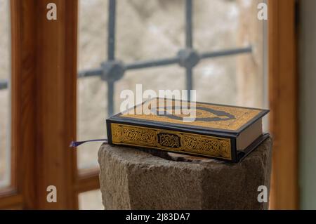 Nahaufnahme des Quran-Buches mit schwarz-goldener Einfassung auf einem Steinsockel neben einem hellen Fenster Stockfoto