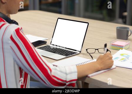 Mittelteil der biracial weibliche Berater schriftlich in Tagebuch von leerem Bildschirm Laptop mit Kopieplatz Stockfoto