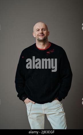 Vertikale mittellange Aufnahme eines selbstbewussten, gut aussehenden, reifen kaukasischen Mannes, der vor grauem Wandhintergrund steht und die Kamera anschaut, mit einem Upcycling-Modekonzept Stockfoto
