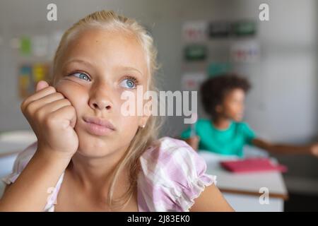 Nahaufnahme eines kaukasischen Grundschulmädchen mit der Hand auf dem Kinn, das wegschaut, während es am Schreibtisch sitzt Stockfoto