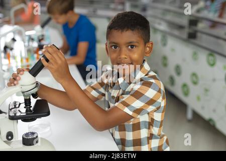 Porträt eines birazialen Grundschuljungen, der das Mikroskop hält, während er im Labor am Schreibtisch sitzt Stockfoto