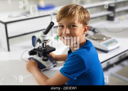 Porträt eines lächelnden kaukasischen Grundschuljungen mit Mikroskop, der im Labor am Schreibtisch sitzt Stockfoto