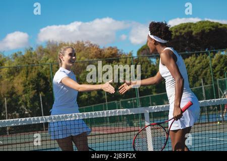 Glückliche, multirassische Tennisspielerinnen, die am sonnigen Tag auf dem Platz die Hände über das Netz schüttelten Stockfoto