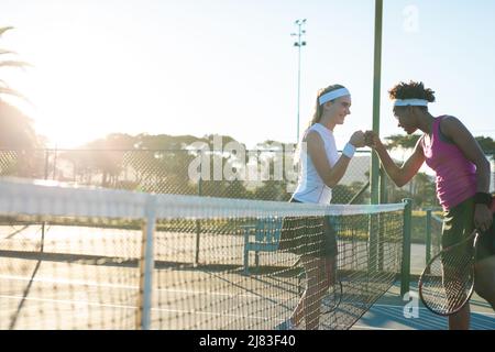 Glückliche, multirassische junge Tennisspielerinnen, die auf dem Platz einen Fauststoß über das Netz geben Stockfoto