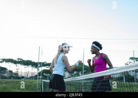 Glückliche, multirassische junge weibliche Spieler, die auf dem Tennisplatz einen Fauststoß über das Netz geben Stockfoto