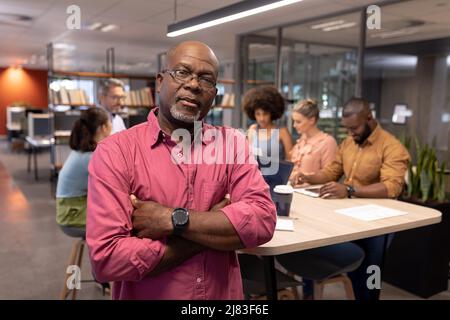 Porträt eines selbstbewussten, reifen afroamerikanischen Geschäftsmannes mit gekreuzten Armen gegen Kollegen Stockfoto