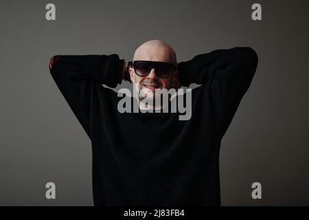 Horizontales mittleres Porträt eines fröhlichen, gut aussehenden, reifen kaukasischen Mannes mit stilvollem Pullover und Sonnenbrille, der vor der Kamera lächelt Stockfoto