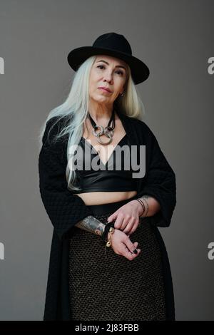 Vertikal mittel lang Studio-Porträt von attraktiven reifen kaukasischen Frau mit langen grauen Haaren tragen moderne Goth Outfit Blick auf die Kamera Stockfoto