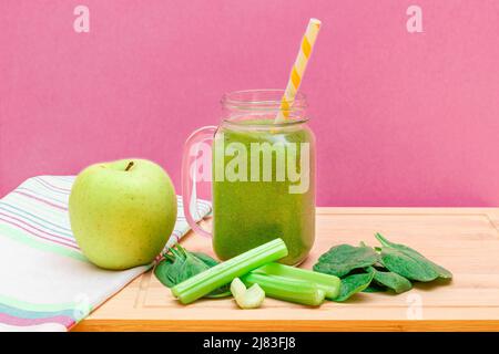 Frischer grüner Smoothie von Apfel, Sellerie und Spinat im gläsernen Smoothie-Glas mit gelbem Cocktailstroh auf Holzschneidebrett. Veganer Detox-Drink. Vegetarische Kultur. Gesunde Ernährung und Obst-Diät Stockfoto