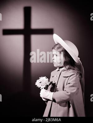 1960S KLEINES MÄDCHEN IN IHREM OSTERSONNTAGSKLEID UND HAUBE MIT EINIGEN GÄNSEBLÜMCHEN MIT CHRISTLICHEM KREUZ IM HINTERGRUND - J11735 HAR001 HARS CHRISTIAN SYMBOLS SPIRITUALITY BONNET B&W GÄNSEBLÜMCHEN VERKNETET UND IM KONZEPT KONZEPTIONELL STILSICHER EINIGE SYMBOLISCHE KONZEPTE KOOPERATION JUGENDLICHE GLAUBEN SCHWARZ-WEISS KAUKASISCHE ETHNIZITÄT HAR001 ALTMODISCHE DARSTELLUNG Stockfoto