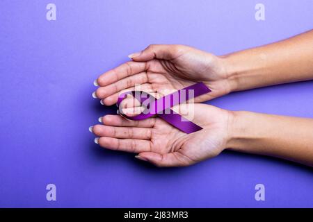 Beschnittene Hände einer afroamerikanischen Frau mit mittlerem Erwachsenen mit violettem Bewusstseinsband auf blauem Hintergrund Stockfoto